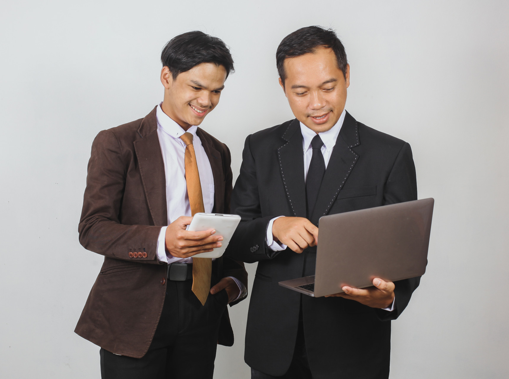 Two smiling businessmen in suits, one holding a tablet and the other using a laptop, engaged in discussion over digital work