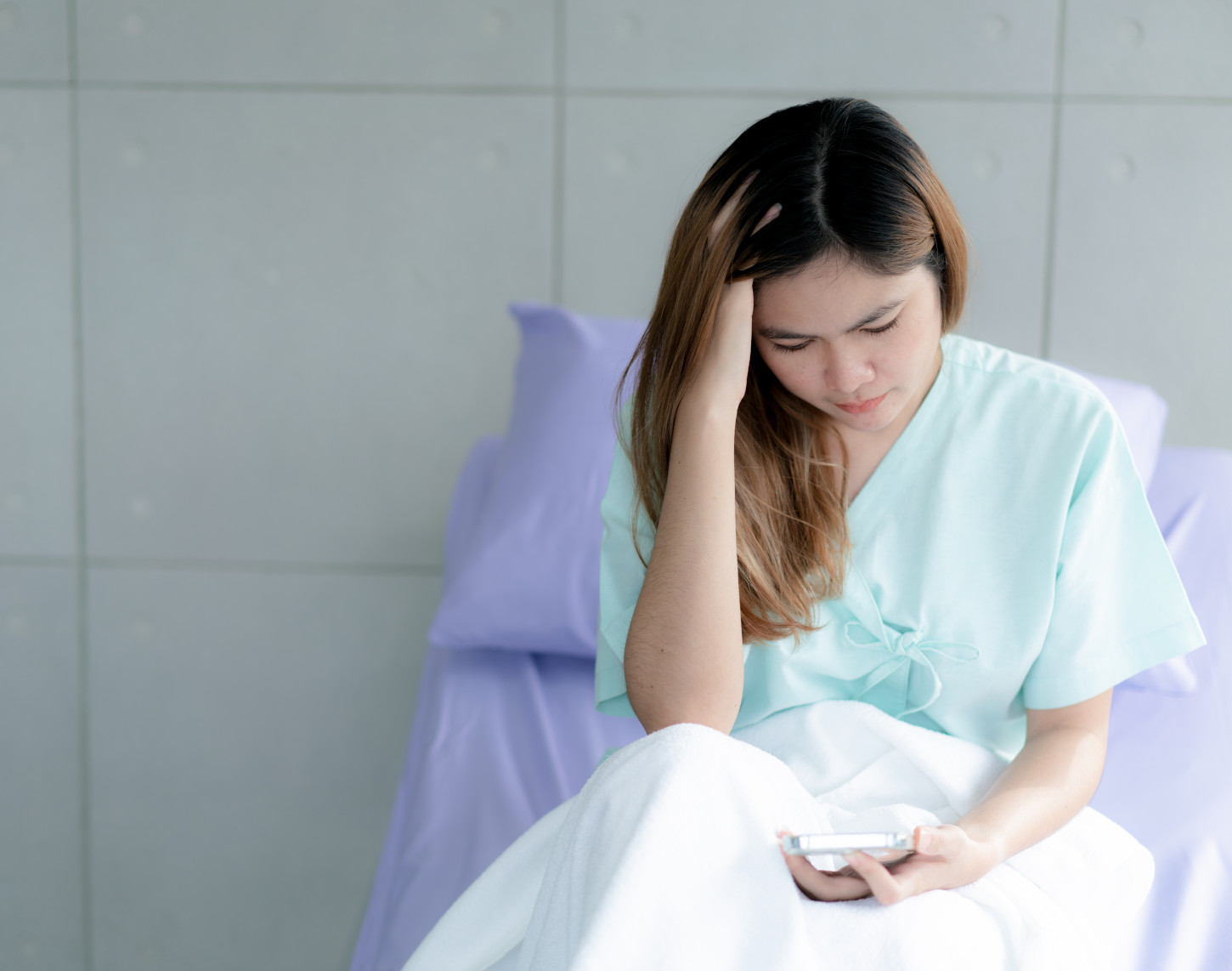 Worried young woman in hospital gown sitting on a bed, looking at a smartphone