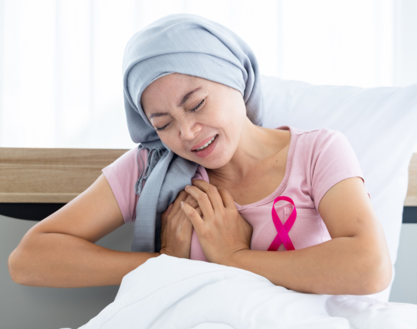 Distressed woman in a headscarf with a pink ribbon, symbolizing breast cancer awareness, clutches her shoulder