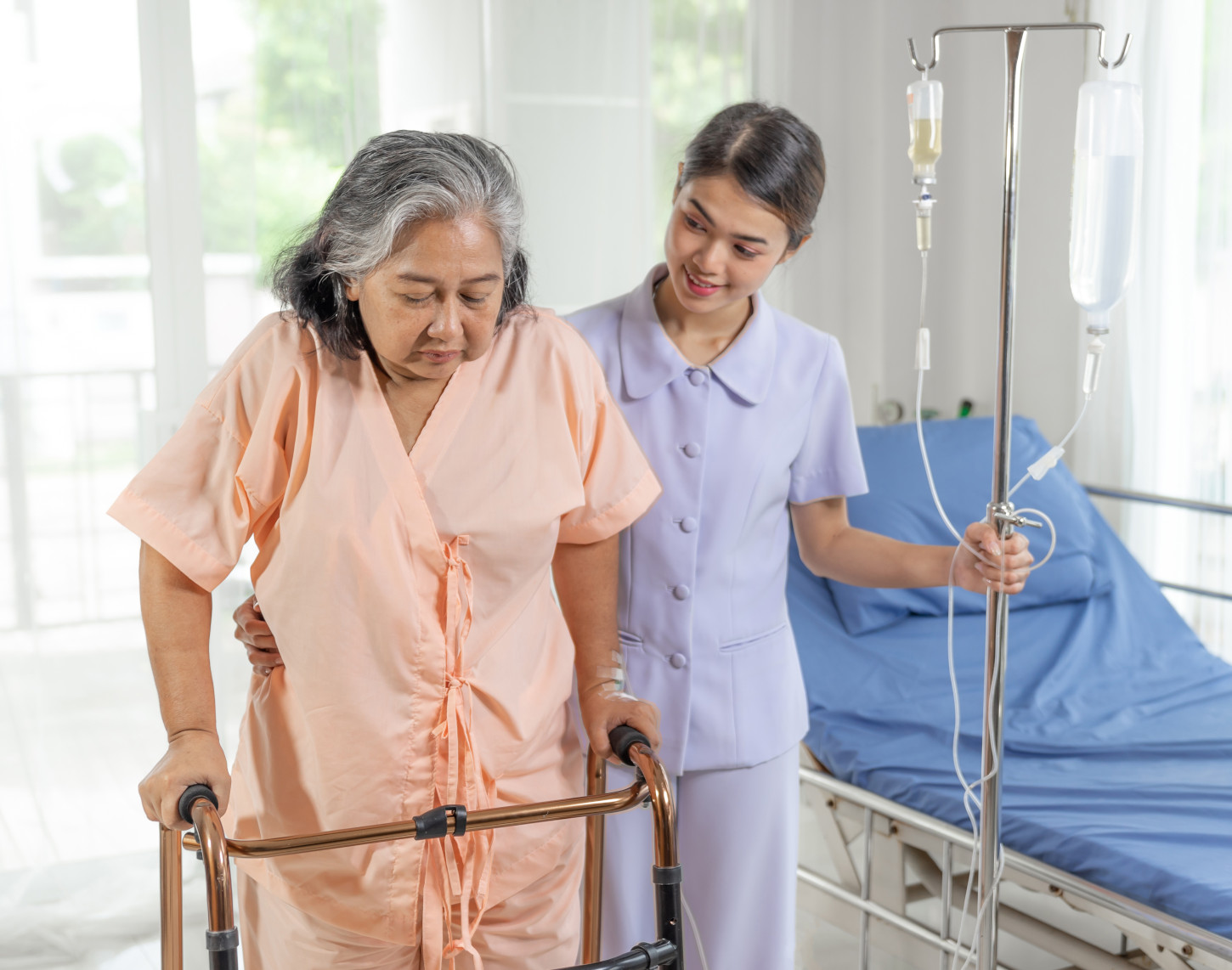 Elderly woman with a walker being assisted by a nurse in a hospital room with an IV stand