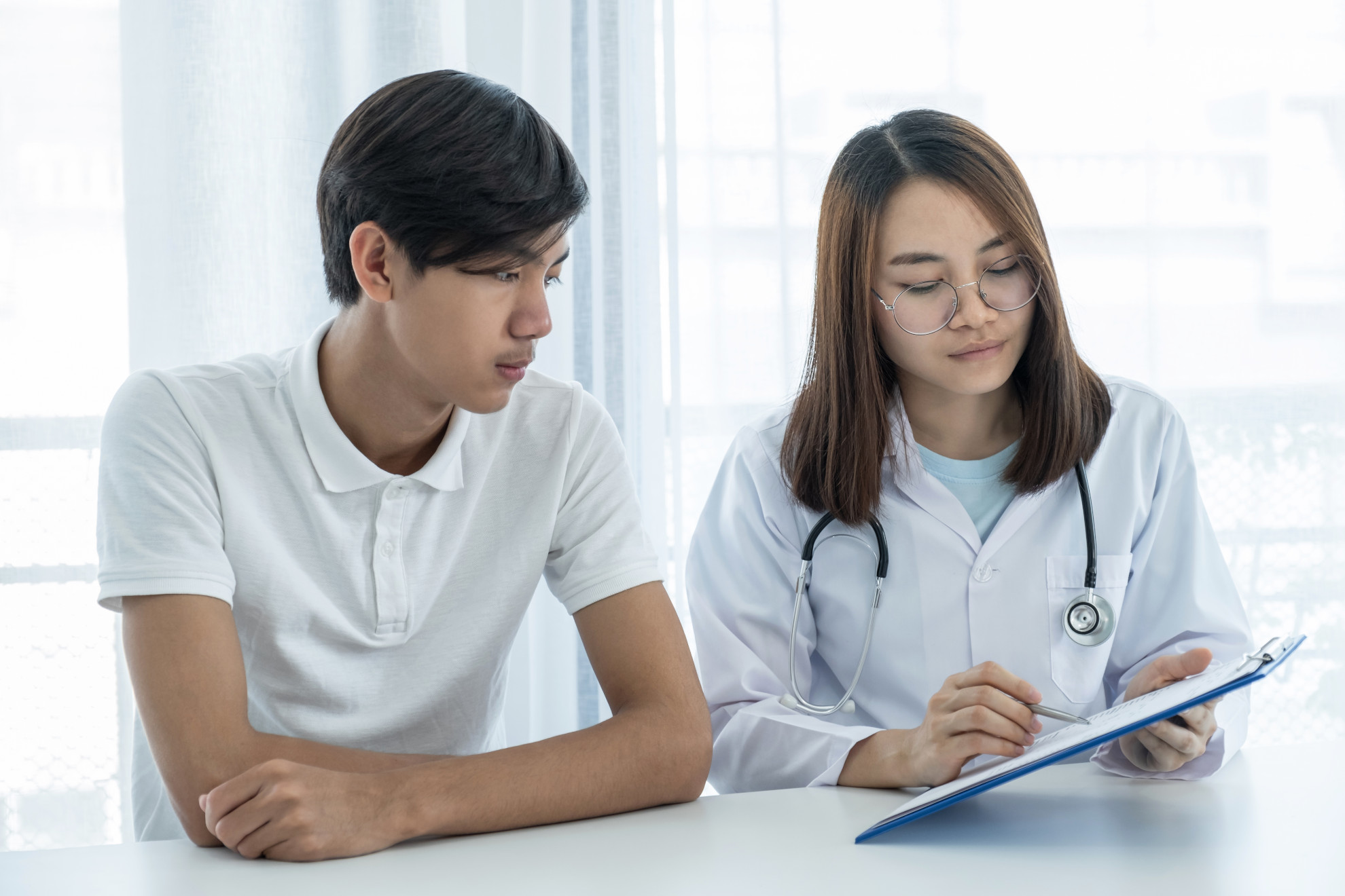 Young male patient with a concerned look sitting next to a female doctor with glasses who is taking notes